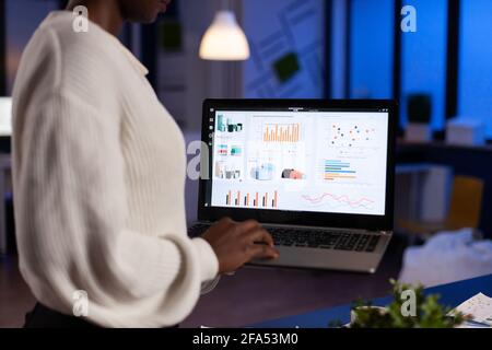 Back view of african business woman analysing financial statistics holding laptop late at night. Busy focused employee using modern technology network wireless overtime. Stock Photo