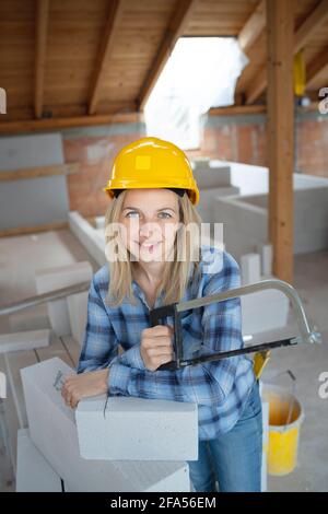 pretty young female bricklayer with yellow safety helmet is sawing bricks on a construction site in the house and is happy Stock Photo