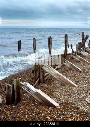 The skeletal ruins of a breakwater with waves crashing over it on a shingle beach under a clouded sky. Stock Photo