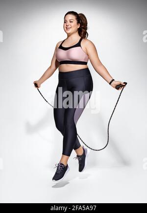 Sporty girl with skipping rope in motion. Photo of model with curvy figure in fashionable sportswear on grey background. Dynamic movement. Sports moti