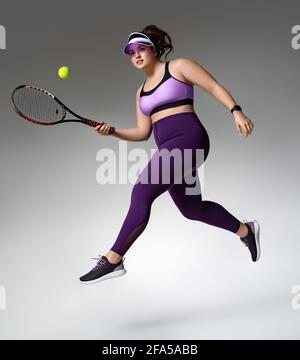 Sporty girl jumping with tennis racquet and ball. Photo of model with curvy figure in fashionable sportswear on grey background. Dynamic movement. Sid Stock Photo