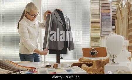 woman dressmaker working with measure tape and tailoring mannequin, adjusting the jacket. Her studio is bright and sunny, clothes and colorful fabrics Stock Photo