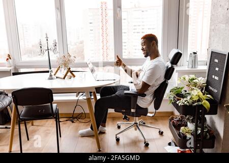 Happy african american businessman is sitting at the desk, using laptop, working from home office. Successful male freelancer having a video meeting Stock Photo