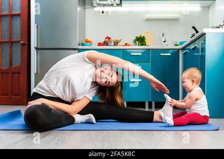 A mother and her baby are stretching on a mat in the kitchen. The child sits near the mother's leg and holds it with his hands. The concept of home sp Stock Photo