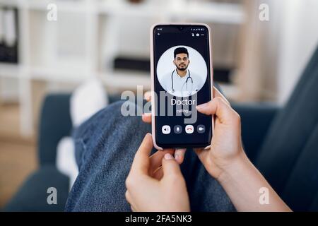 Close-up back view of female patient hands, using smart phone and calling to her doctor, young Indian man. App for remote medical service. Stock Photo