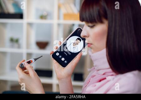 Close up side view of young sick woman, suffering from a respiratory illness, sore throat and fever, making a video call to male Indian gp doctor. Online consultation. Stock Photo