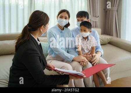 Asian woman's insurance broker is offering details of health insurance coverage for COVID-19 to Asian family with father, mother and son in their livi Stock Photo