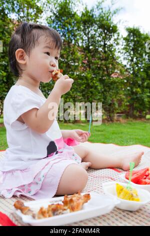 Adorable little asian (thai) girl enjoy eating her lunch in the park, child eating fried chicken, chinese steamed dumpling and watermelon over blur gr Stock Photo