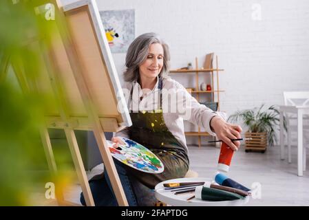 mature woman holding paintbrush and palette while reaching red paint in tube and sitting near canvas Stock Photo