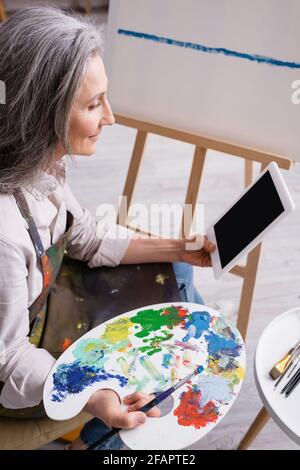mature woman holding palette and digital tablet with blank screen near canvas Stock Photo