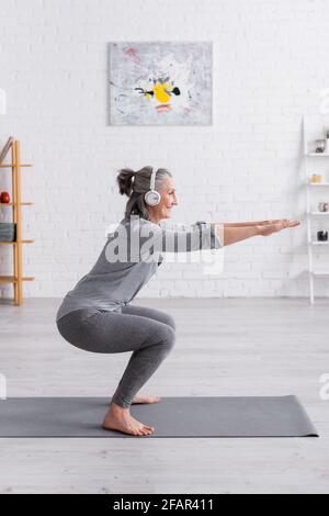 side view of cheerful middle aged woman in sportwear and headphones doing squat on mat at home Stock Photo