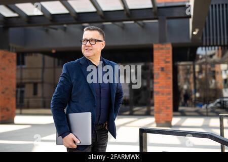 Handsome mature businessman wearing stylish eyeglasses holding laptop computer, standing on the street. Portrait of smiling developer looking at camer Stock Photo