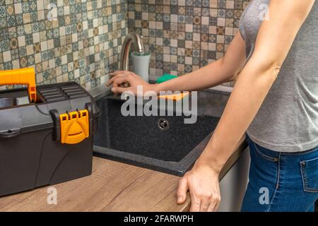 Young woman stands by the faucet and thinks about fixing it herself in a modern kitchen, next to a toolbox Stock Photo