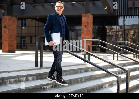 Handsome mature businessman wearing stylish eyeglasses holding laptop computer, standing on the street. Portrait of smiling developer looking at camer Stock Photo