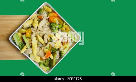 Young corn capcay vegetables with mushrooms and also peppers are seen from the above on a white box plate against a green background Stock Photo