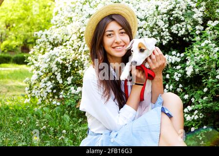 Portrait of attractive brunette young woman with cute jack russell terrier puppy, green lawn, foliage, flowers, park background. Female in hipster str Stock Photo