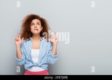 worried woman in plaid shirt looking up while holding crossed fingers on grey background Stock Photo