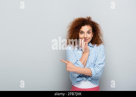 surprised woman pointing with finger while looking at camera on grey background Stock Photo