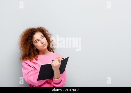 pensive woman holding notebook and looking away on grey background Stock Photo