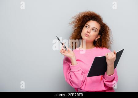 thoughtful woman looking away while holding pen and notebook on grey background Stock Photo