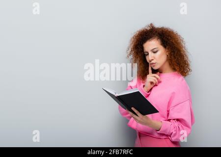 curly woman holding notebook and touching face while thinking on grey background Stock Photo
