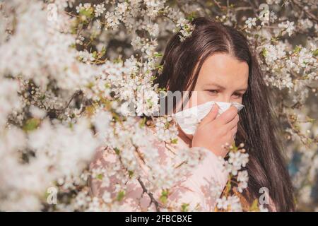 Spring allergy symptoms concept, young woman sneezing in front of blooming a tree, allergy to pollen and flowering season, healthcare Stock Photo