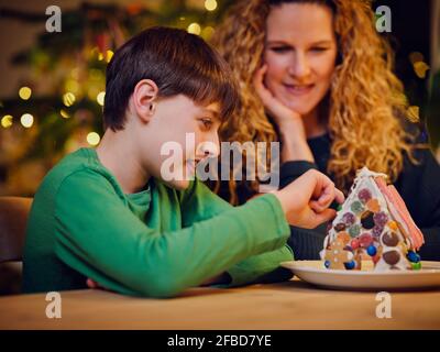Smiling cute boy decorating gingerbread house by mother during Christmas Stock Photo