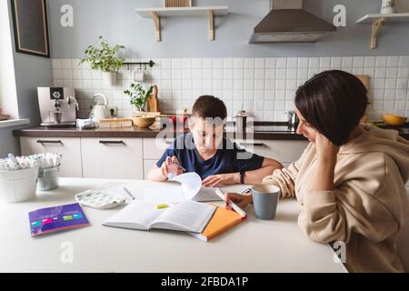 Mother assisting son in homework while sitting at dining table in kitchen at home Stock Photo
