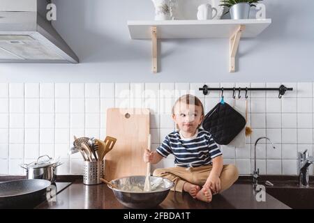 Playful toddler with ladle and bowl looking away while sitting on kitchen counter at home Stock Photo