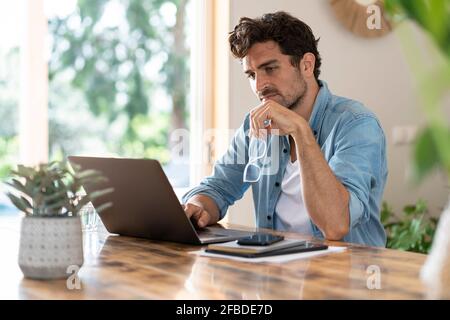 Concentrated male freelancer holding eyeglasses while working on laptop at table Stock Photo