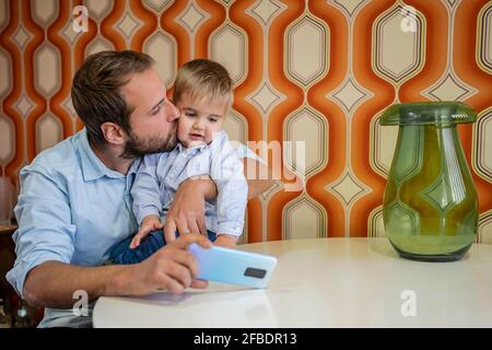 Caring father kissing son while taking selfie at table Stock Photo