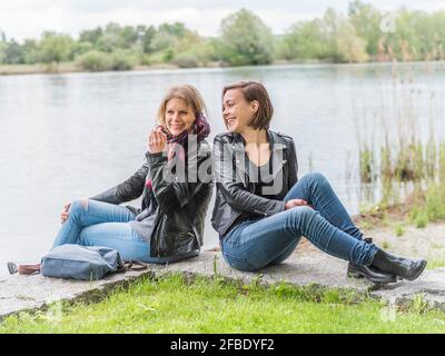 Two smiling friends sitting near water having fun Stock Photo