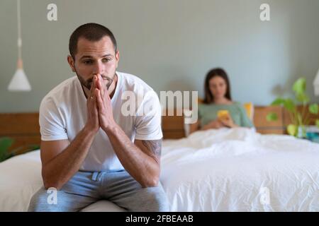 Worried young man sitting on bed while girlfriend using smart phone in background at home Stock Photo