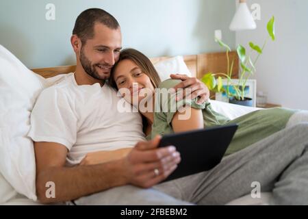 Smiling young couple using digital tablet while lying on bed at home Stock Photo