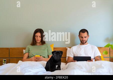 Couple using smart phone and digital tablet while sitting on bed with Pug dog in bedroom at home