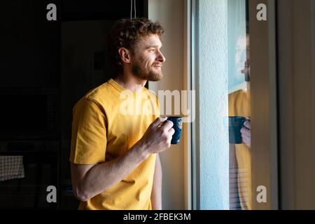 Smiling man looking through window while having coffee at home Stock Photo