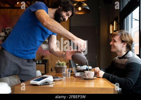 Male entrepreneur pouring hot water from kettle to coworker sitting at office Stock Photo