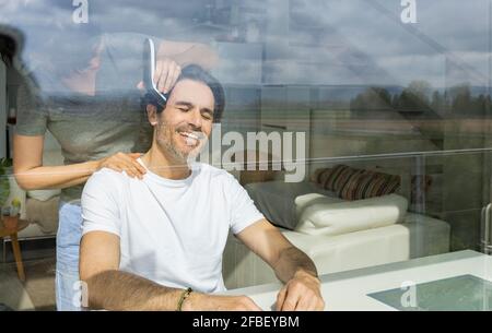 Woman cutting man's hair using electric razor while sitting at home seen through glass Stock Photo