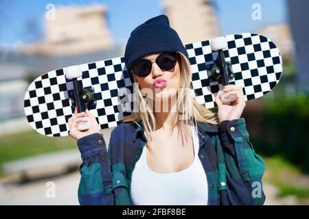 Young woman in knit hat puckering while carrying skateboard on shoulder during sunny day Stock Photo