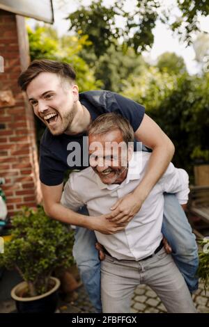 Happy father giving piggyback ride to laughing son in backyard Stock Photo