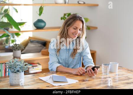 Smiling blond female entrepreneur surfing net through smart phone while sitting at table Stock Photo