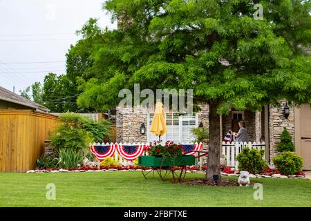 Cute rock house decorated for 4th of July bunting and flags with nice landscaping and a huge walnut tree and woman on porch unfolding more decorations Stock Photo