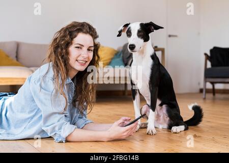 Pretty woman lying on wooden floor using a smartphone with dog sitting next to her both looking into camera Stock Photo