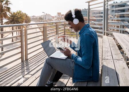 Cheerful man pointing using smart phone over laptop while sitting on wooden steps at promenade Stock Photo