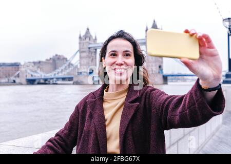 Smiling young woman taking selfie with Tower Bridge through smart phone Stock Photo