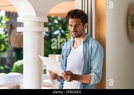 Serious man reading document while leaning on doorframe at home Stock Photo