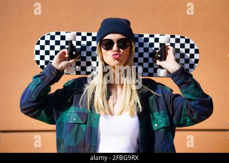 Blond woman puckering while carrying skateboard on shoulder in front of brown wall Stock Photo
