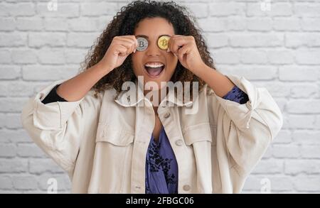 Excited curly haired woman covering eyes with gold and silver colored bitcoins in front of wall Stock Photo