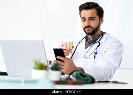 Handsome male healthcare worker drinking coffee while using mobile phone Stock Photo