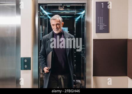 Mature businessman with laptop and luggage walking out of lift in hotel Stock Photo